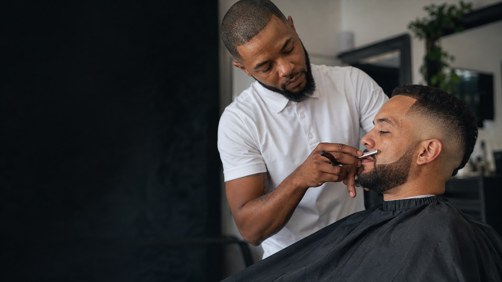 Adult male barber in a white shirt shaping a beard beside a fresh fade